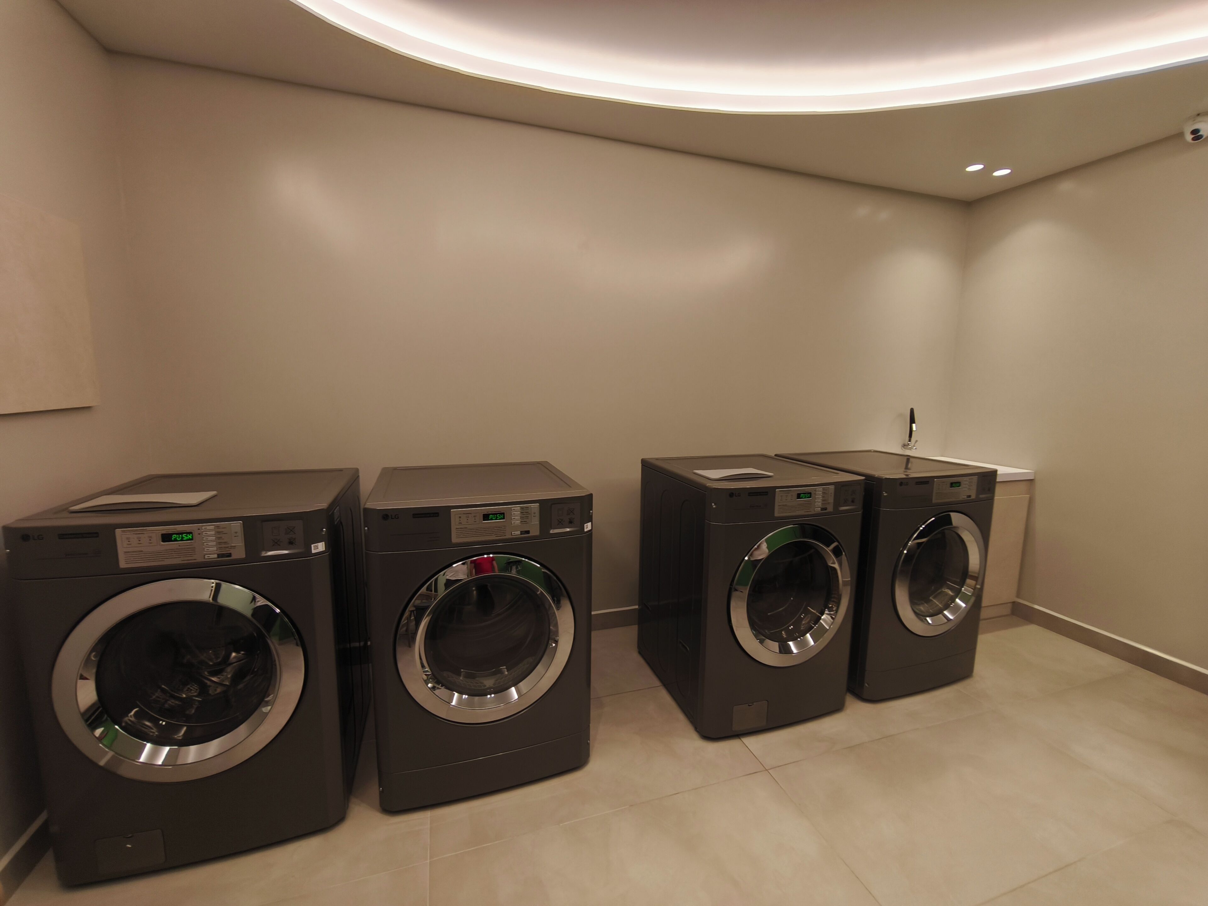 A laundry room with four front-loading washing machines, a wall-mounted dryer, and a ceiling light fixture.