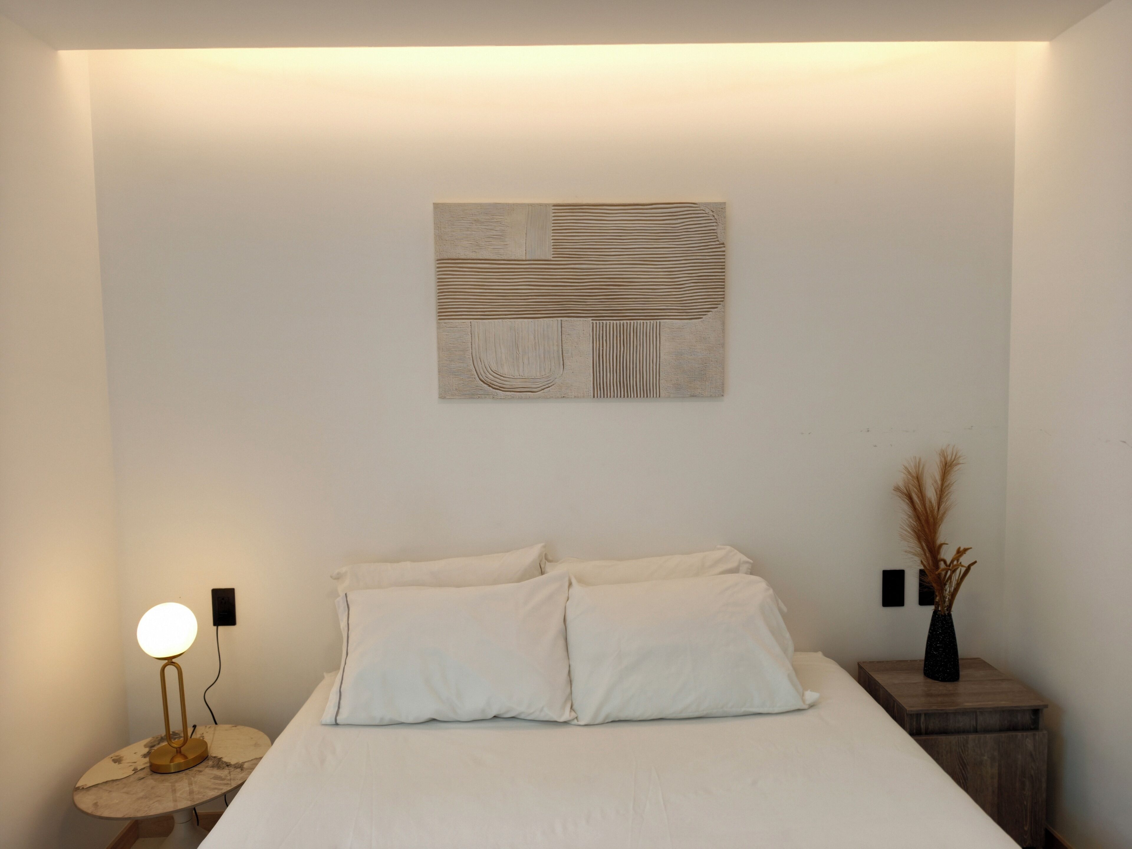 A bedroom with a bed, a wall-mounted abstract art piece, a bedside lamp, and a small table with a vase.
