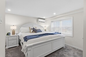 Une chambre avec un lit blanc, deux tables de chevet, une fenêtre avec des stores et un appareil de climatisation.