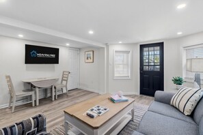 A modern living room with a sofa, a coffee table, a TV mounted on the wall, a dining table with chairs, and a door. - North End 1BR/1BA Unit 4 - Walk to Beach & Boardwalk (Ocean City)