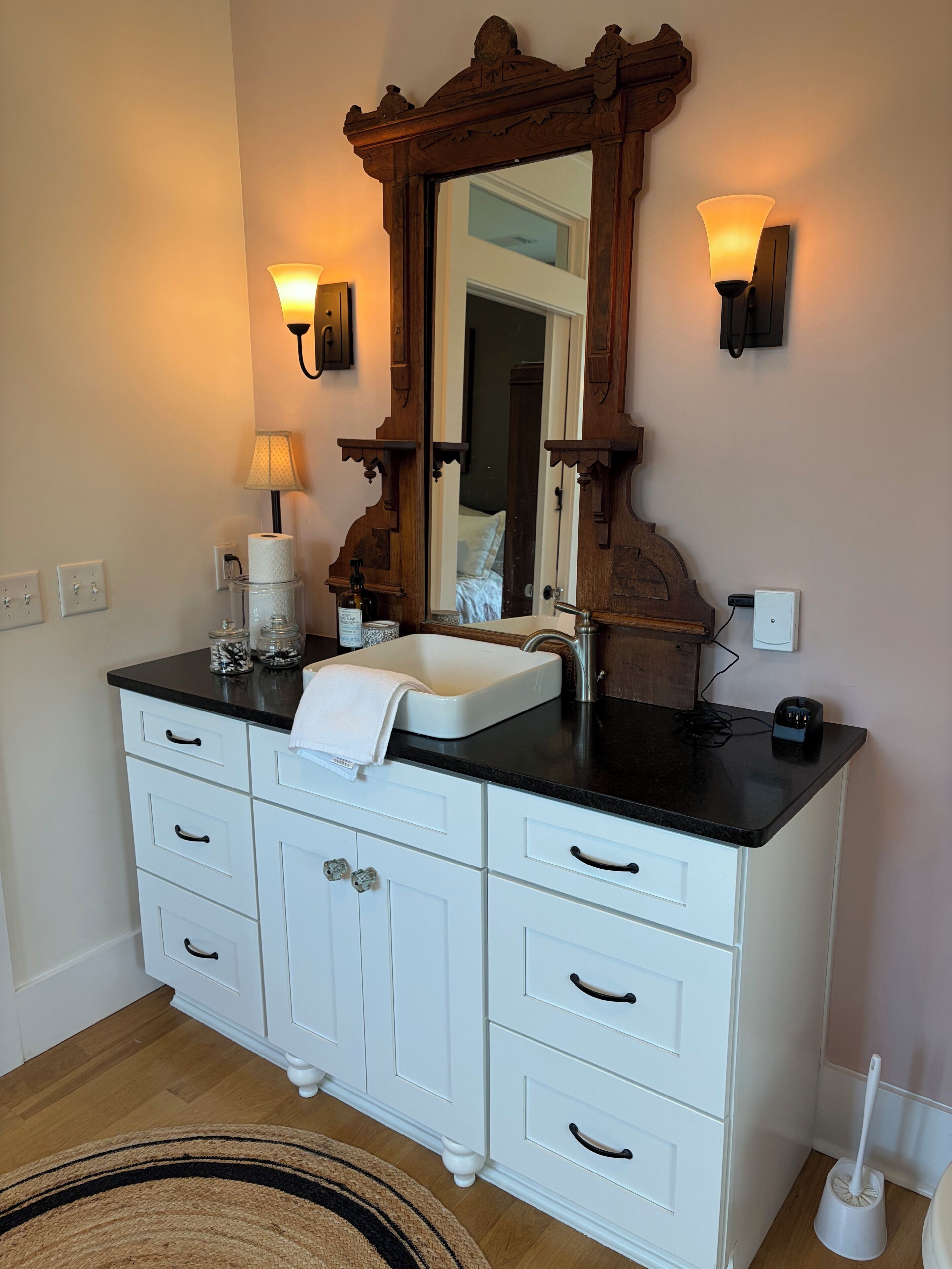 A bathroom with a white vanity, a mirror with an ornate frame, two wall-mounted lights, a towel on the sink, and a floor lamp.