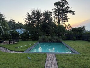 Ein rechteckiger Pool mit einem Backsteinweg, umgeben von einer Rasenfläche und einem Holzzaun.