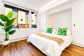 Een slaapkamer met een groot bed, een raam met uitzicht op gebouwen en een kamerplant.