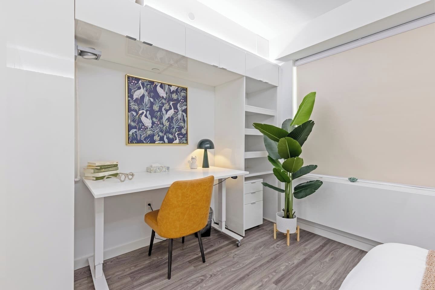 A modern bedroom with a white desk, a yellow chair, a large potted plant, and a bed with a white headboard.