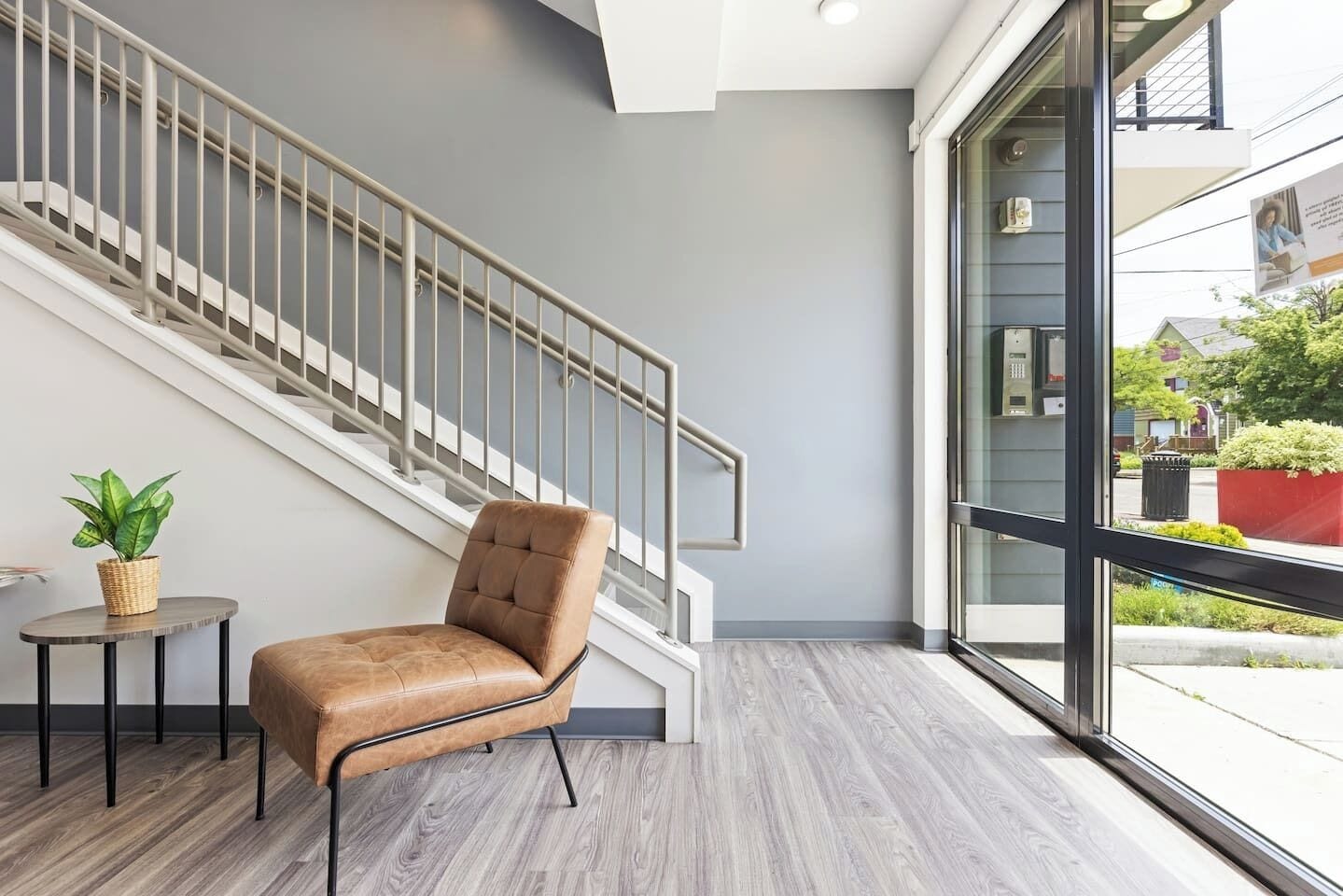 Un interior moderno con una escalera, un sillón de cuero, una mesita con una planta y un ventanal que ofrece vistas al exterior.