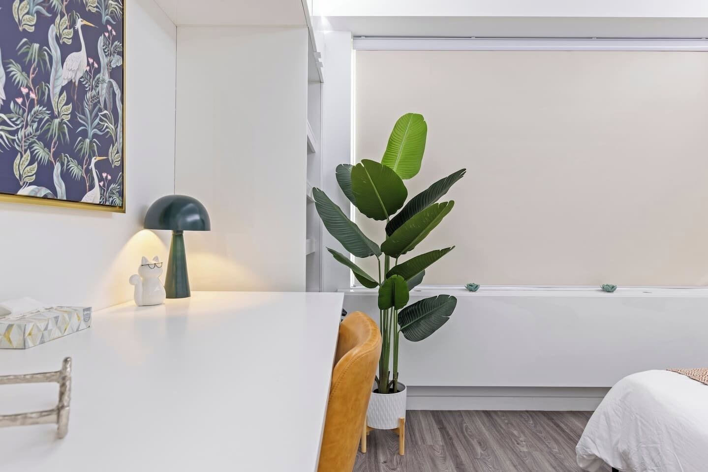 Une chambre moderne avec un bureau blanc, une lampe, une plante et un lit recouvert d’une couverture blanche.