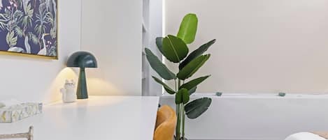 Una habitación moderna con un escritorio blanco, una lámpara, una planta y una cama con cubrecama blanco.