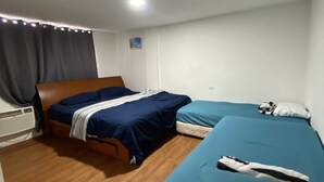Ein Zimmer mit zwei Betten, eines mit einem Holzkopfende und das andere mit einem weißen Kopfende. Es gibt einen kleinen Tisch mit einer Lampe, ein Fenster mit Vorhängen und einen Wandheizkörper.