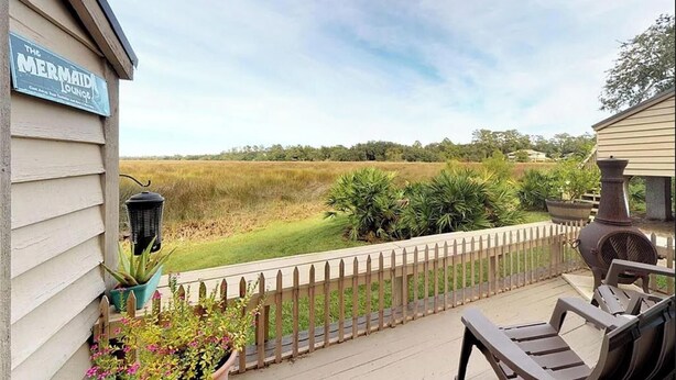 A deck with a view of a grassy field, a wooden fence, and a sign for 'The Mermaid Lounge'. - Moss & Marsh | Marshfront Views, Pool & Island Charm (Saint Simons Island)