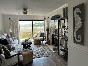 A living room with a sofa, ottoman, and a fireplace. There is a view of a field through the window. - Moss & Marsh | Marshfront Views, Pool & Island Charm (Saint Simons Island)