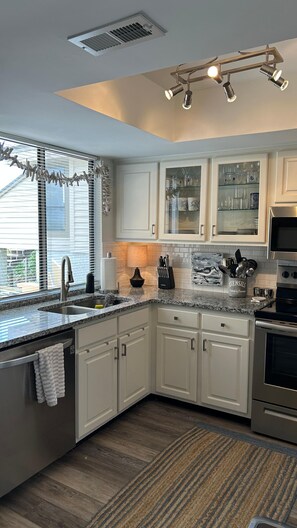 A modern kitchen with white cabinets, a granite countertop, a built-in oven, and a window with blinds. - Moss & Marsh | Marshfront Views, Pool & Island Charm (Saint Simons Island)