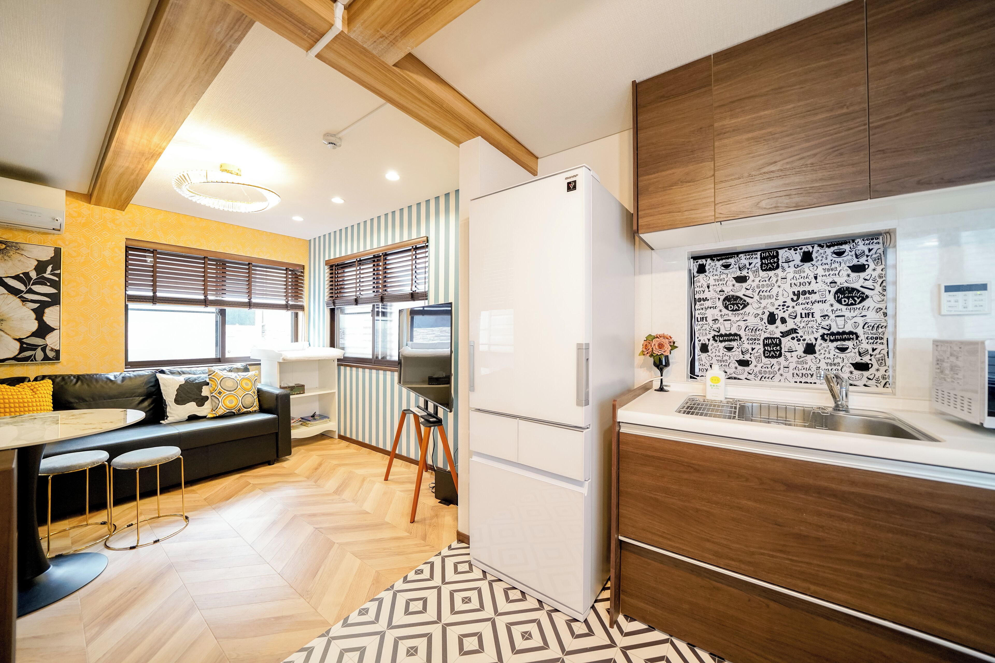 A modern kitchen with a refrigerator, wooden cabinets, a dining area with a sofa and table, and a patterned floor.