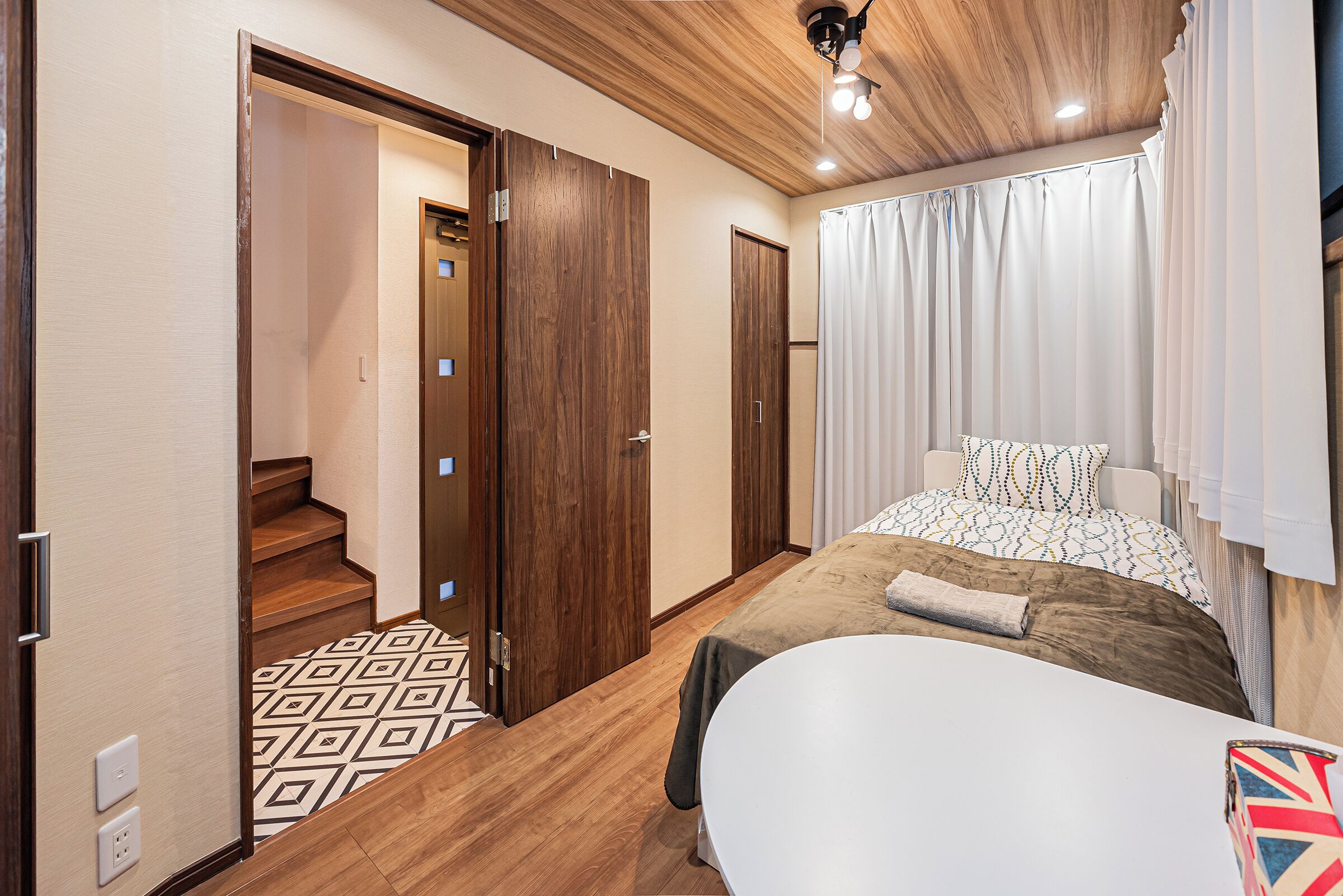 A bedroom with a bed, a bedside table, a window with curtains, a wooden ceiling, and a staircase leading to another room.