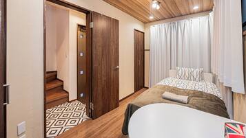 A bedroom with a bed, a bedside table, a window with curtains, a wooden ceiling, and a staircase leading to another room.