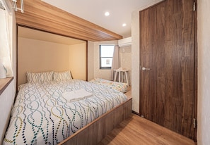 A bedroom with a wooden headboard, a bed with a patterned bedspread, a small stool, and a window with curtains.