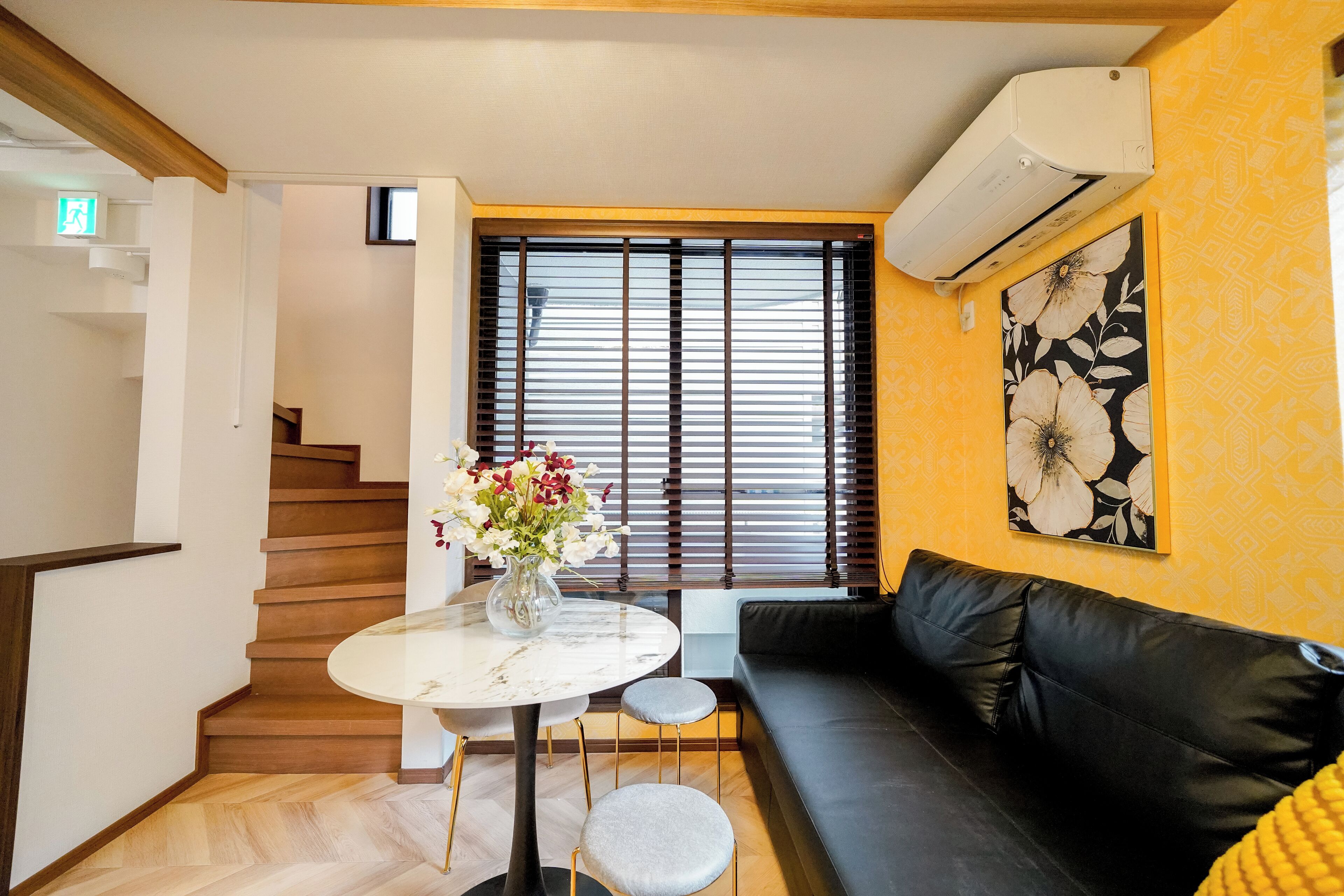 A modern living room with a black leather sofa, a round marble table with a vase of flowers, a staircase with wooden steps, and a large window with blinds.