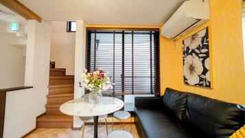 A modern living room with a black leather sofa, a round marble table with a vase of flowers, a staircase with wooden steps, and a large window with blinds.