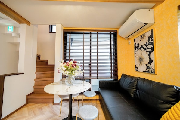 A modern living room with a black leather sofa, a round marble table with a vase of flowers, a staircase with wooden steps, and a large window with blinds.