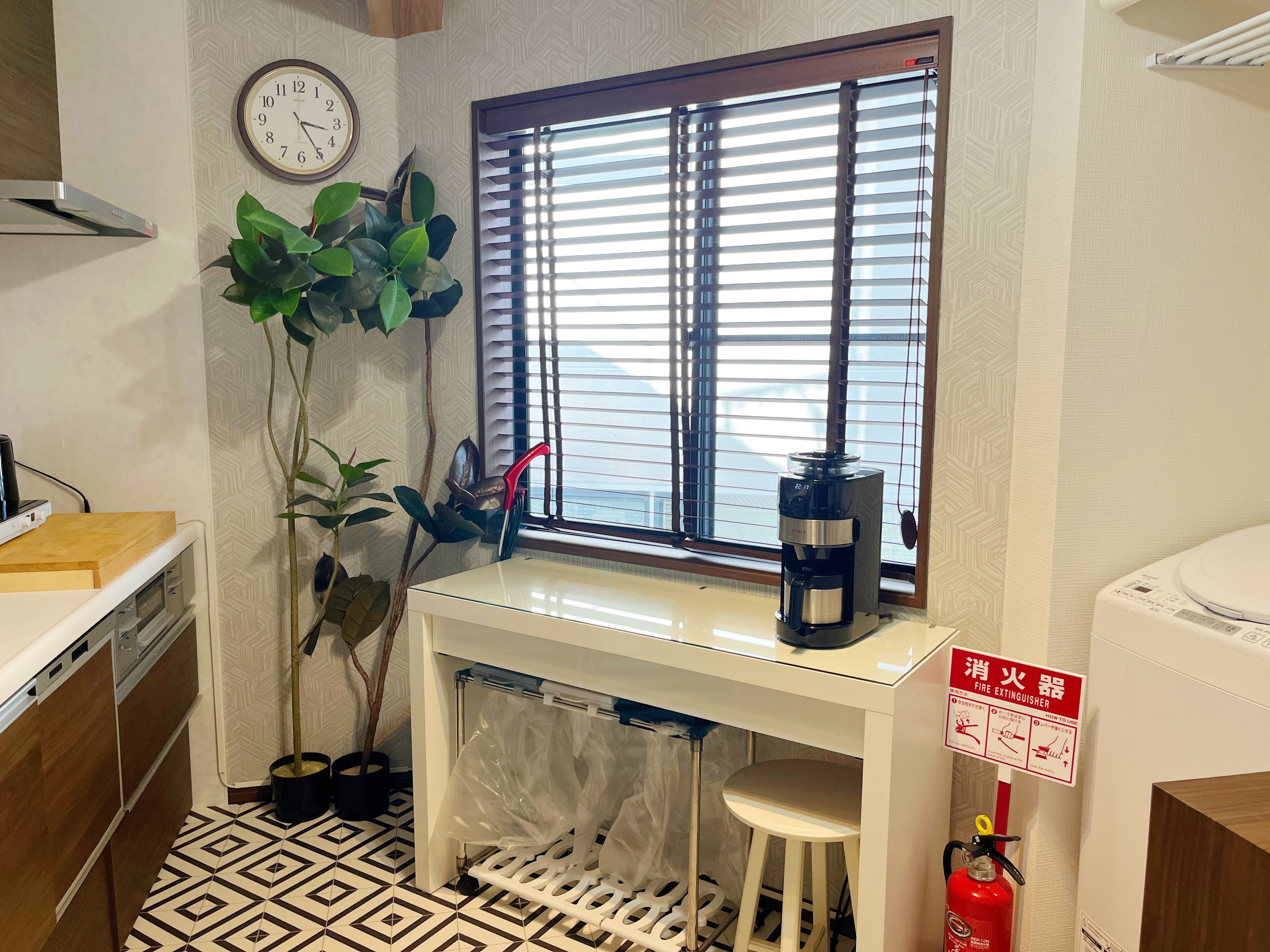 A compact kitchen with a window, a clock, a coffee maker, a fire extinguisher, and a stool.