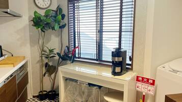 A compact kitchen with a window, a clock, a coffee maker, a fire extinguisher, and a stool.