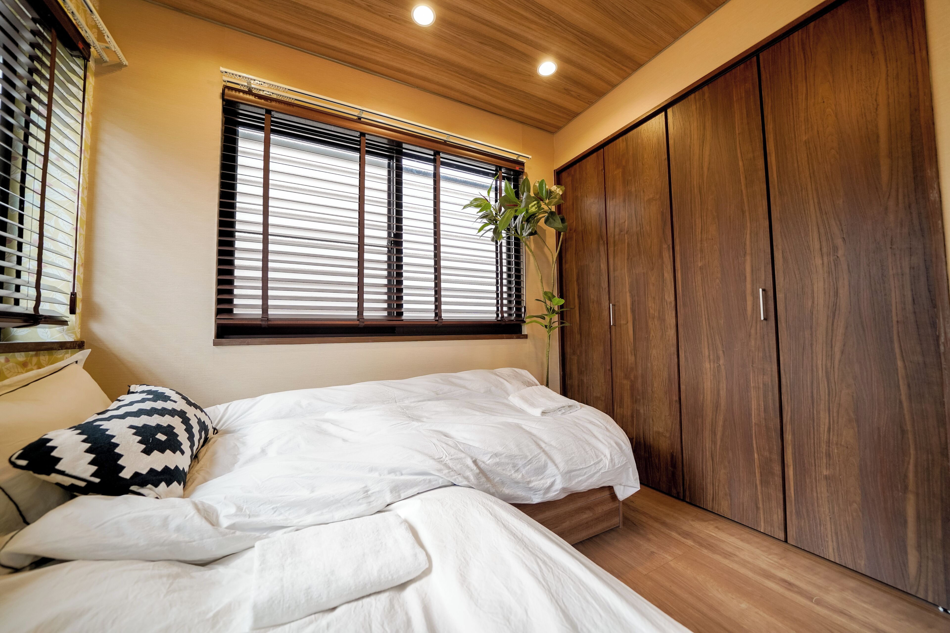 A bedroom with a bed, a window with blinds, a wardrobe, and a plant.