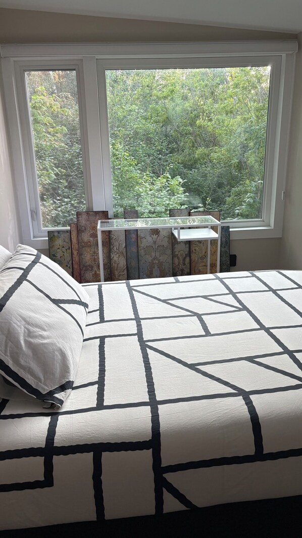 A bedroom with a large window, a bed with a geometric-patterned bedspread, and a series of decorative panels on the window sill.