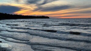 Un paysage côtier avec des vagues, une rive bordée d’une forêt et un ciel au coucher du soleil.