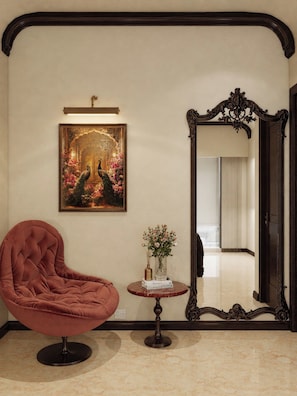 A room with a large mirror, a framed painting, a plush chair, and a small table with a vase.