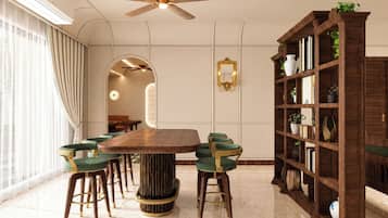 Een eetgedeelte met een ronde tafel, groene bekleden stoelen, een hoge houten boekenkast en een plafondventilator.