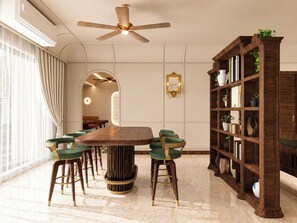 A dining area with a round table, green upholstered chairs, a tall wooden bookshelf, and a ceiling fan.