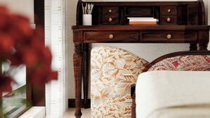 Een kamer met een houten console tafel, een ottoman met patroon, een ingelijst schilderij aan de muur, en een vloerkleed in blauw en beige tinten.