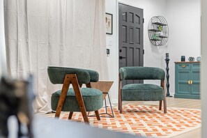 Une pièce comprenant un fauteuil recouvert d’un tissu vert, une chaise en bois et une petite table. On y trouve un tapis à motifs sur le sol, une porte au fond et un meuble vert orné d’objets décoratifs.