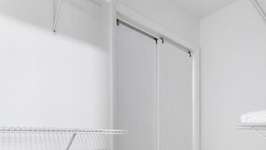 Una habitación con suelo de madera, paredes blancas y techo blanco. Hay un ventilador de techo blanco, un radiador blanco y una estantería blanca.