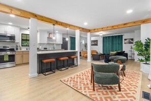 Una cocina moderna con una isla central, taburetes de bar y una zona de comedor con un tapete estampado y una silla tapizada de color verde.