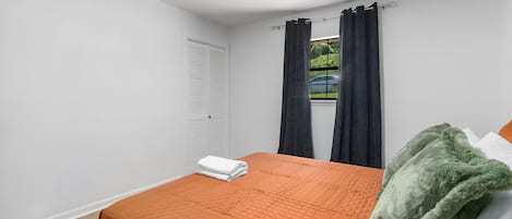 A bedroom with a bed, a window with curtains, a radiator, and a view of the outside.