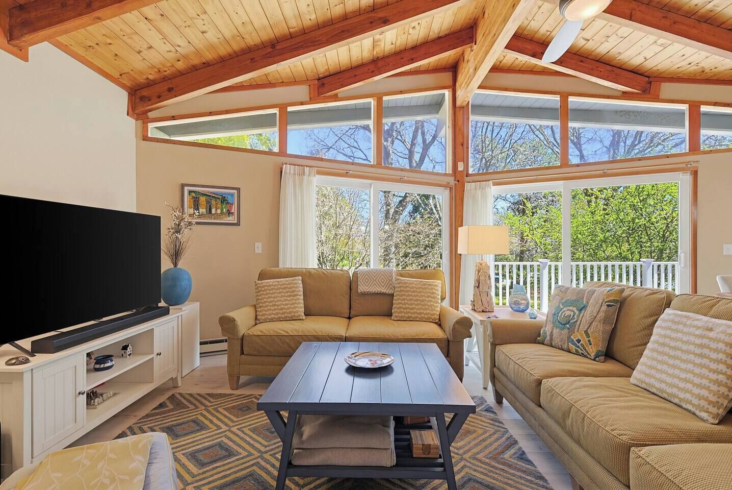 A cozy living room with a large flat-screen TV, a sofa set, a coffee table, and a view through large windows.