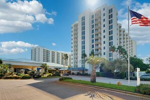 A multi-story hotel with a central courtyard, palm trees, and an American flag. - 2407 Stunning 2 bdrm ~ Book Oct 26th ~ Pool ~ Gym ~ Onsite Restaurant & Bar (Destin)