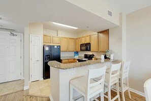 A modern kitchen with granite countertops, wooden cabinets, a black refrigerator, a microwave, and a stove. There are white chairs around a small island. - 2407 New Listing Special ~ Book Sept 7th ~ Pools ~ Gym ~ Onsite Restaurant & Bar (Destin)