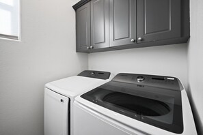 A laundry area with a front-loading washer and dryer, a cabinet above the dryer, and a window to the left. - Heron by Nest Villas | Entertainers Dream Backyard (Indio)