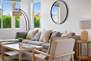 A modern living room with a sectional sofa, a coffee table, a side table with a lamp, and a large mirror on the wall. - Phoebe by Nest Villas | Private Escape w Game Room (Indio)
