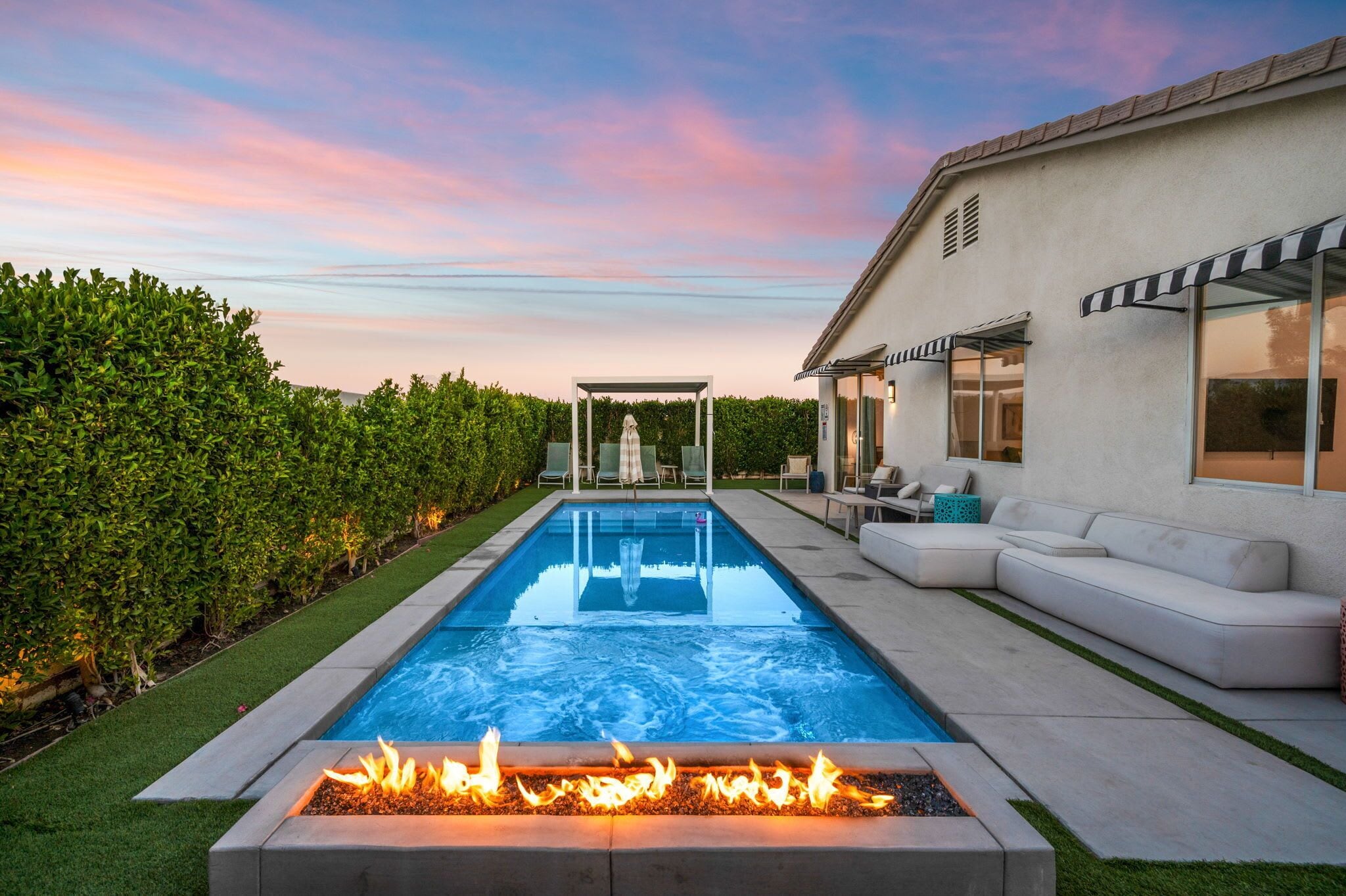A rectangular swimming pool with a fire feature in the foreground, surrounded by a well-maintained hedge and a grassy area.