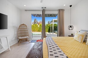 A modern bedroom with a bed, a chair, a TV, and a view of a pool and greenery. - Phoebe by Nest Villas | Private Escape w Game Room (Indio)