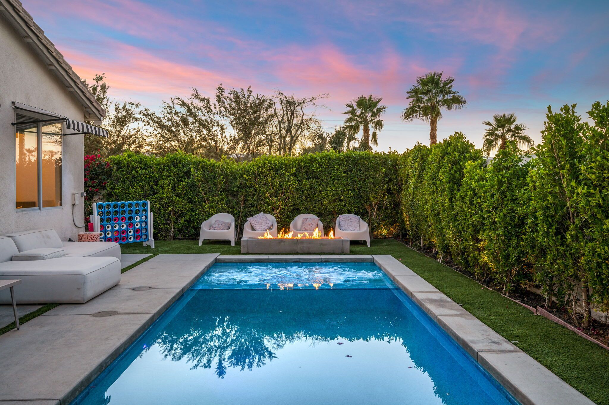 Ein rechteckiger Pool mit Feuerstelle und Sitzgelegenheiten, umgeben von einer Hecke und Palmen.
