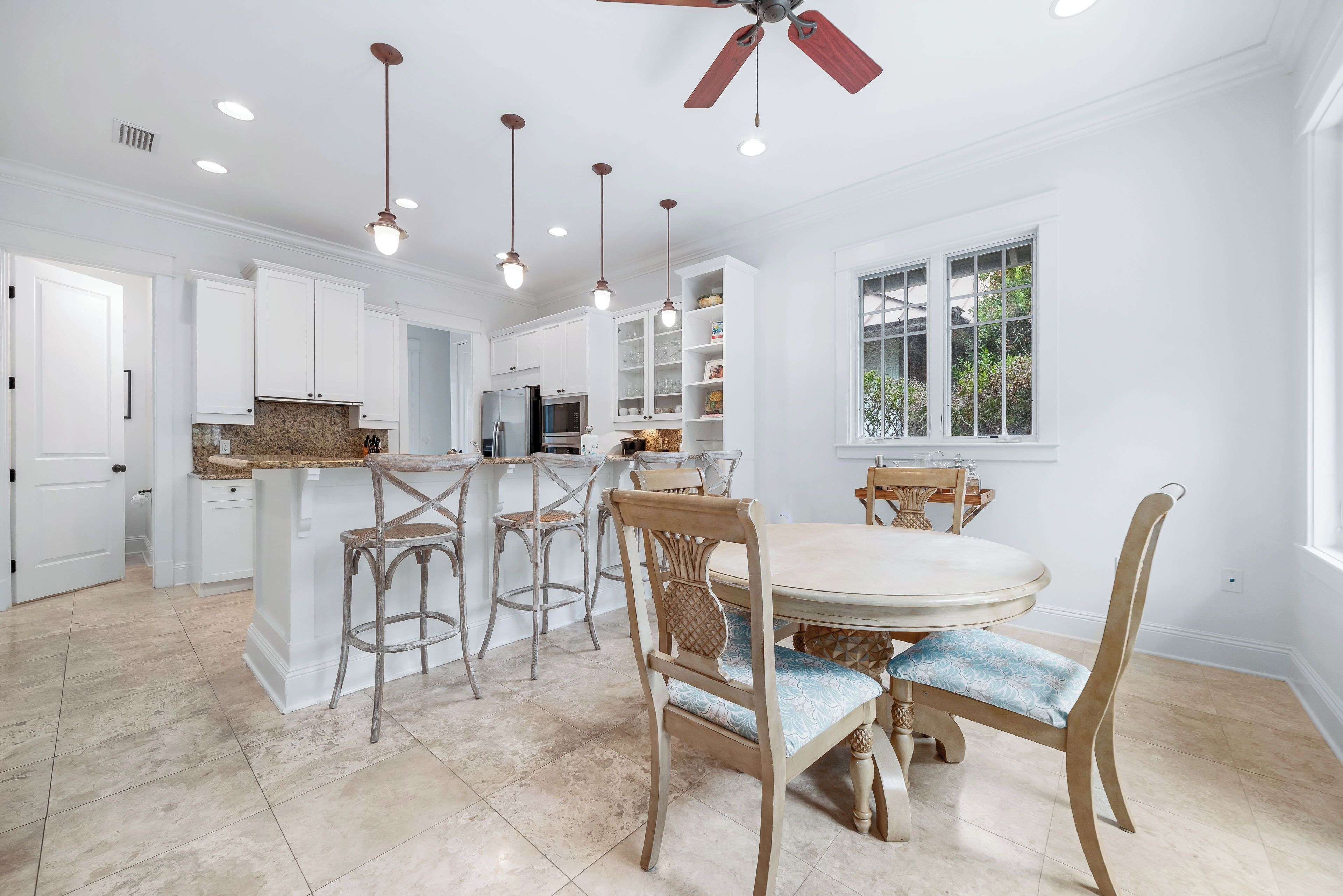 A modern kitchen with white cabinetry, a central island, bar stools, a dining table with chairs, pendant lighting, and a ceiling fan.