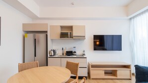 A modern kitchen with a refrigerator, microwave, and stove, a dining area with a round table and wicker chairs, and a living area with a flat-screen TV and a sofa.
