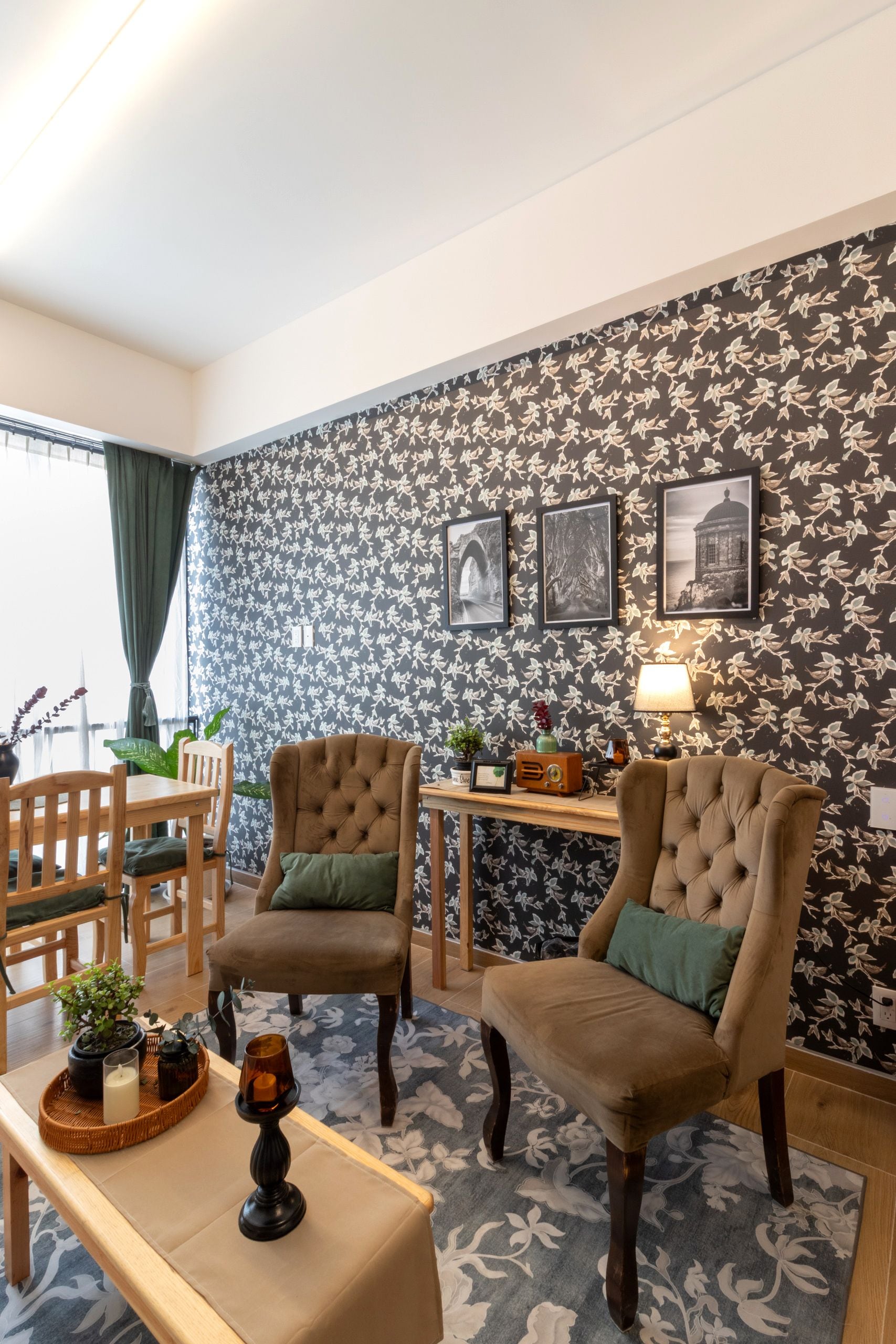 A room with two upholstered chairs, a wooden table, a lamp, and a wall with floral wallpaper and framed pictures.