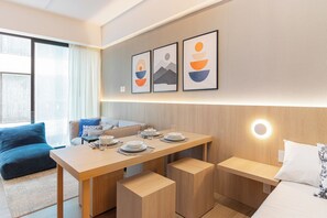 Habitación de hotel moderna con una mesa de comedor de madera, un sofá y una cama. Hay cuadros en la pared y una lámpara de pared.