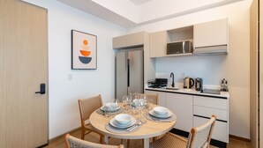 A modern kitchen with white cabinets, a stainless steel refrigerator, and a microwave. There is a dining table with chairs and a framed picture on the wall.