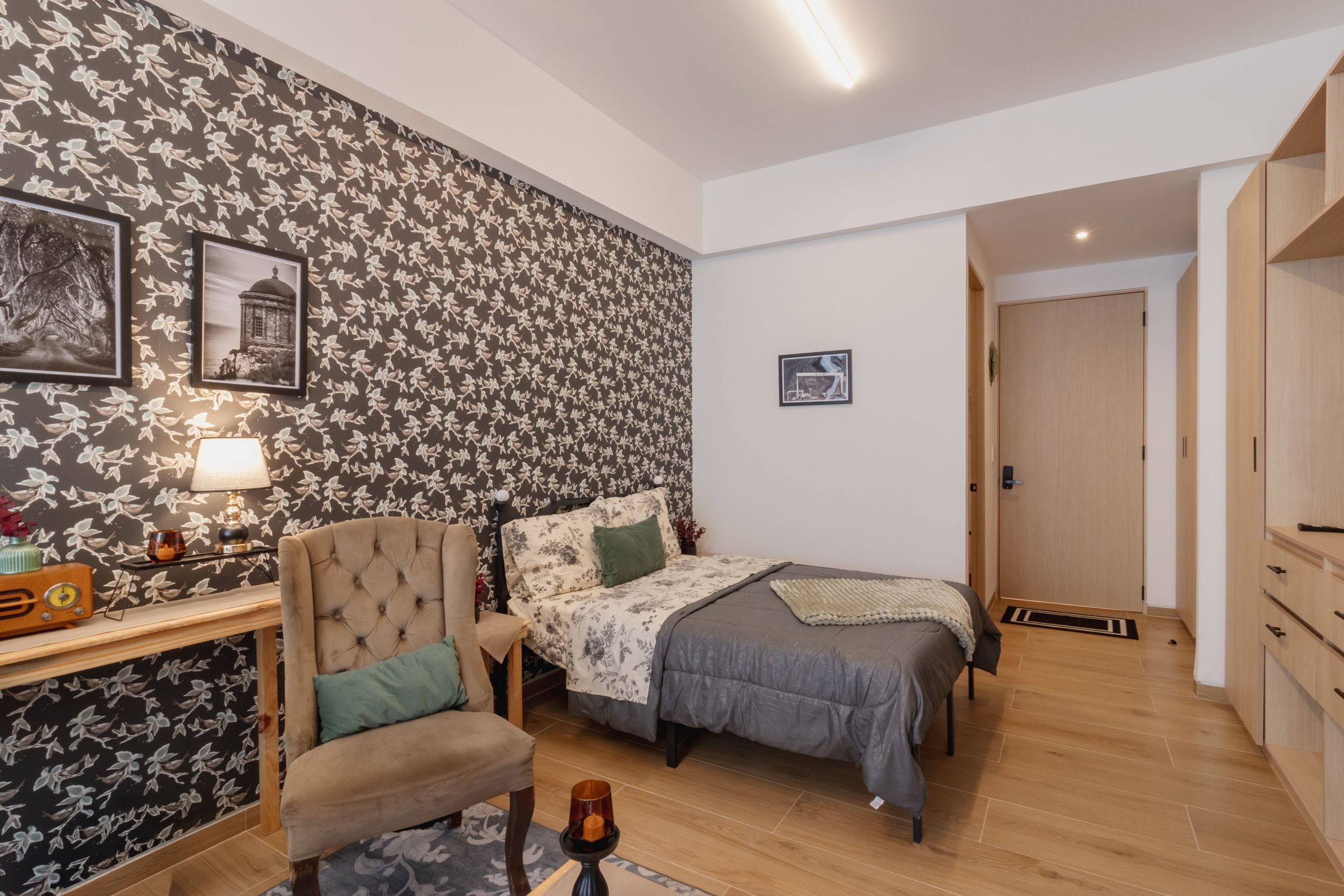 Un dormitorio con cama, una silla, un escritorio con lámpara y un papel tapiz floral en la pared.