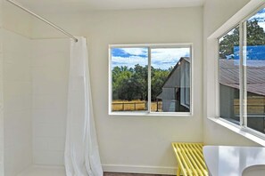 A bathroom with a shower, a window with a view of a building and trees, and a bench. - Maple Ring Kenwood Cottage (Kenwood)